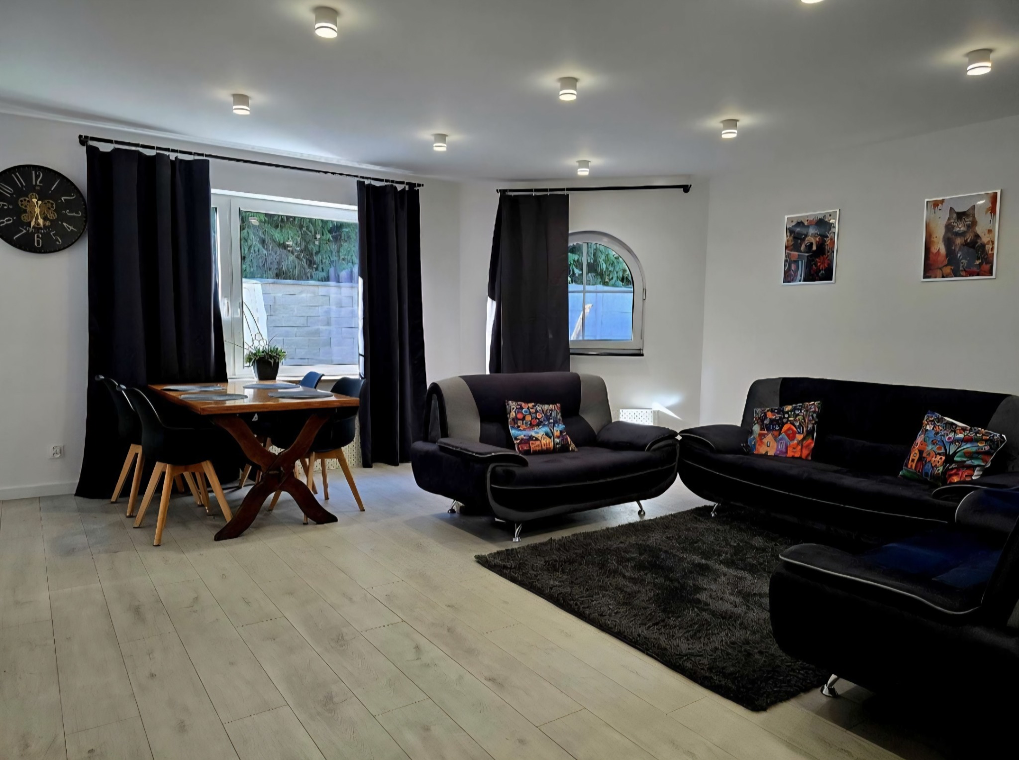 Jasny salon z drewnianym stołem, czarnymi krzesłami i kanapami. Na ścianach obrazy i zegar. Okna z czarnymi zasłonami wpuszczają naturalne światło.