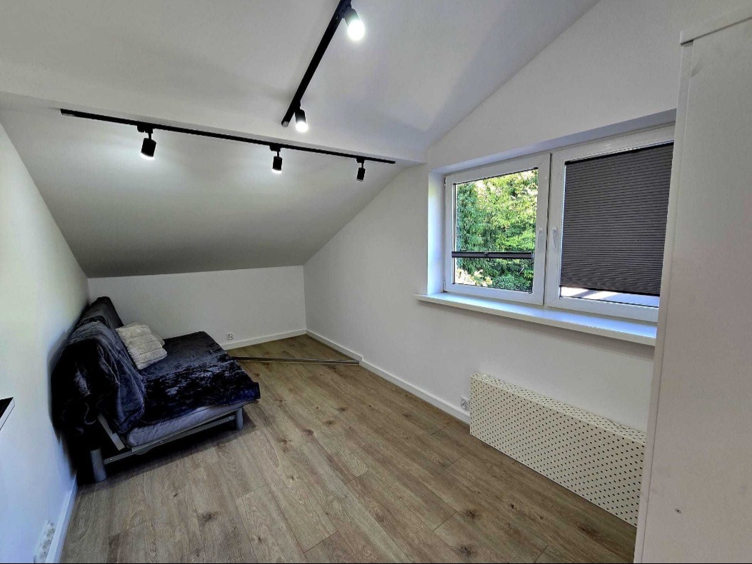 Pokój na poddaszu po remoncie: białe ściany, drewniana podłoga, okno z roletą, szara sofa i nowoczesne oświetlenie szynowe. Skos sufitu dodaje przestrzeni unikalnego charakteru.