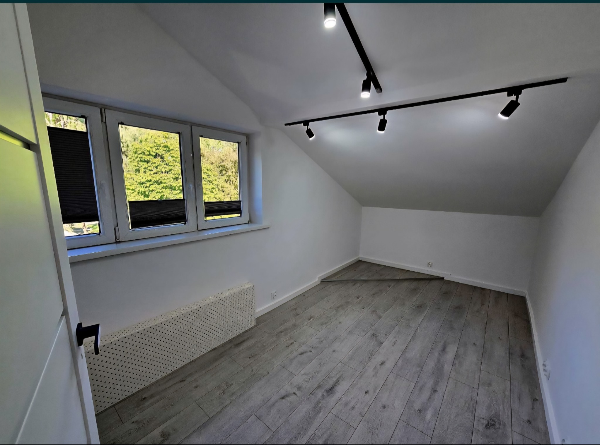 Pokój na poddaszu po remoncie: szare panele, białe ściany, okno z żaluzjami i nowoczesne oświetlenie sufitowe. Minimalistyczny design.