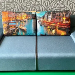 Niebieska sofa z poduszkami w weneckie krajobrazy na tle wzorzystej tapety. Widok z przodu na ustawiony mebel.