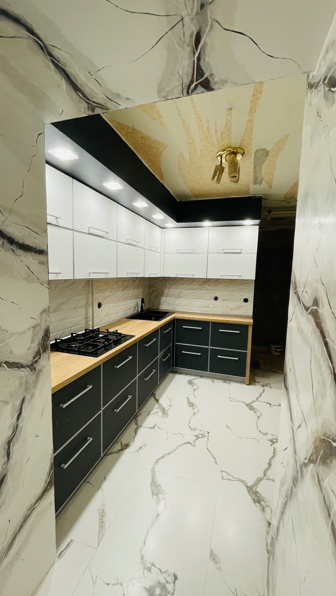 Kompaktowa, narożna kuchnia z czarno-białymi szafkami i drewnianym blatem. Marmurowe płytki na podłodze i ścianach. Widok z perspektywy wejścia.