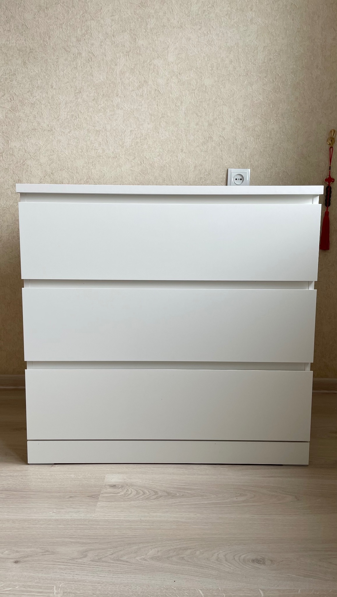 Biała komoda z trzema szufladami na jasnej podłodze, widoczny fragment ściany z gniazdkiem i dekoracyjnym elementem. Minimalistyczny design.