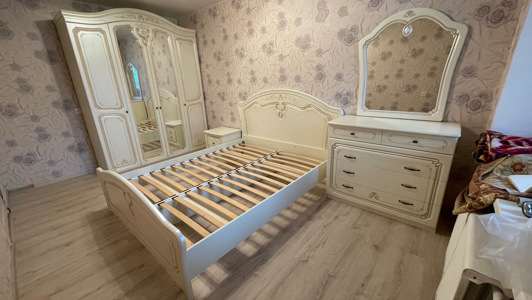 Komplet mebli do sypialni w kremowym kolorze z dekoracyjnymi zdobieniami: łóżko bez materaca, szafa z lustrami i komoda z lustrem. Wzór tapety w tle.