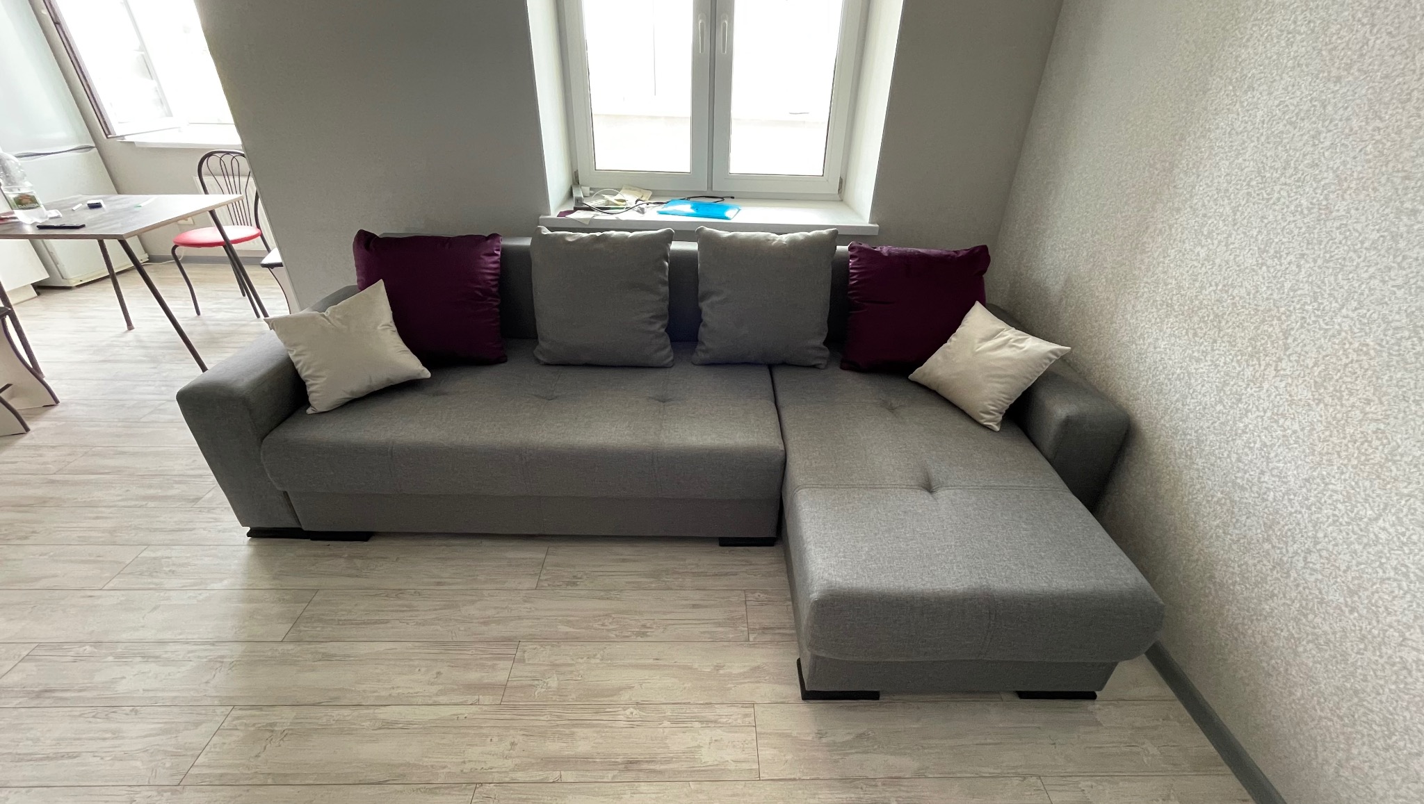 Szary narożnik z poduszkami w pokoju z oknem. Widoczne panele podłogowe i fragment stołu. Stonowana kolorystyka wnętrza, minimalistyczny styl.