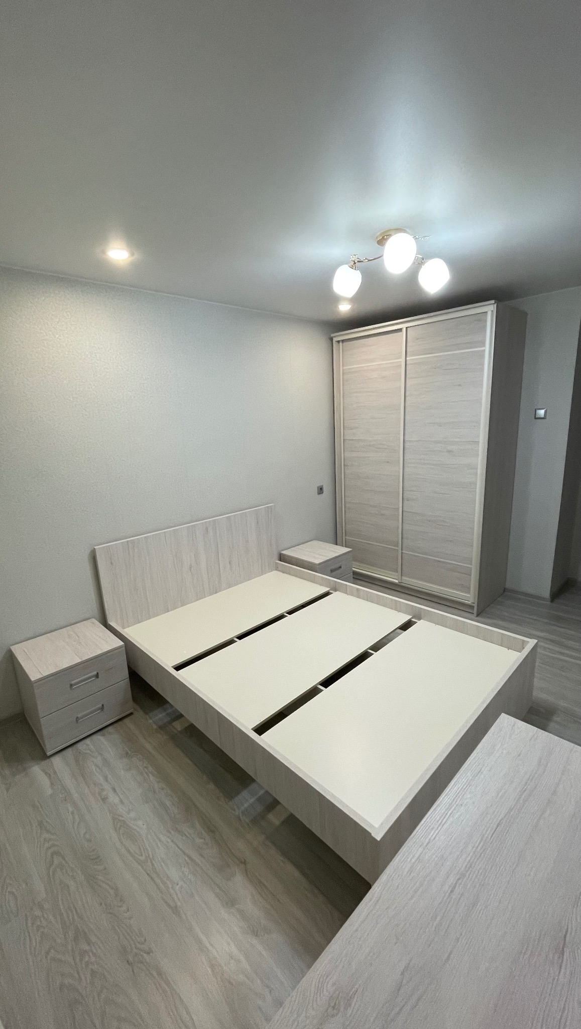 Jasne, drewniane łóżko bez materaca i szafka nocna w minimalistycznym pokoju. Widoczna również szafa przesuwna. Neutralna kolorystyka pomieszczenia.