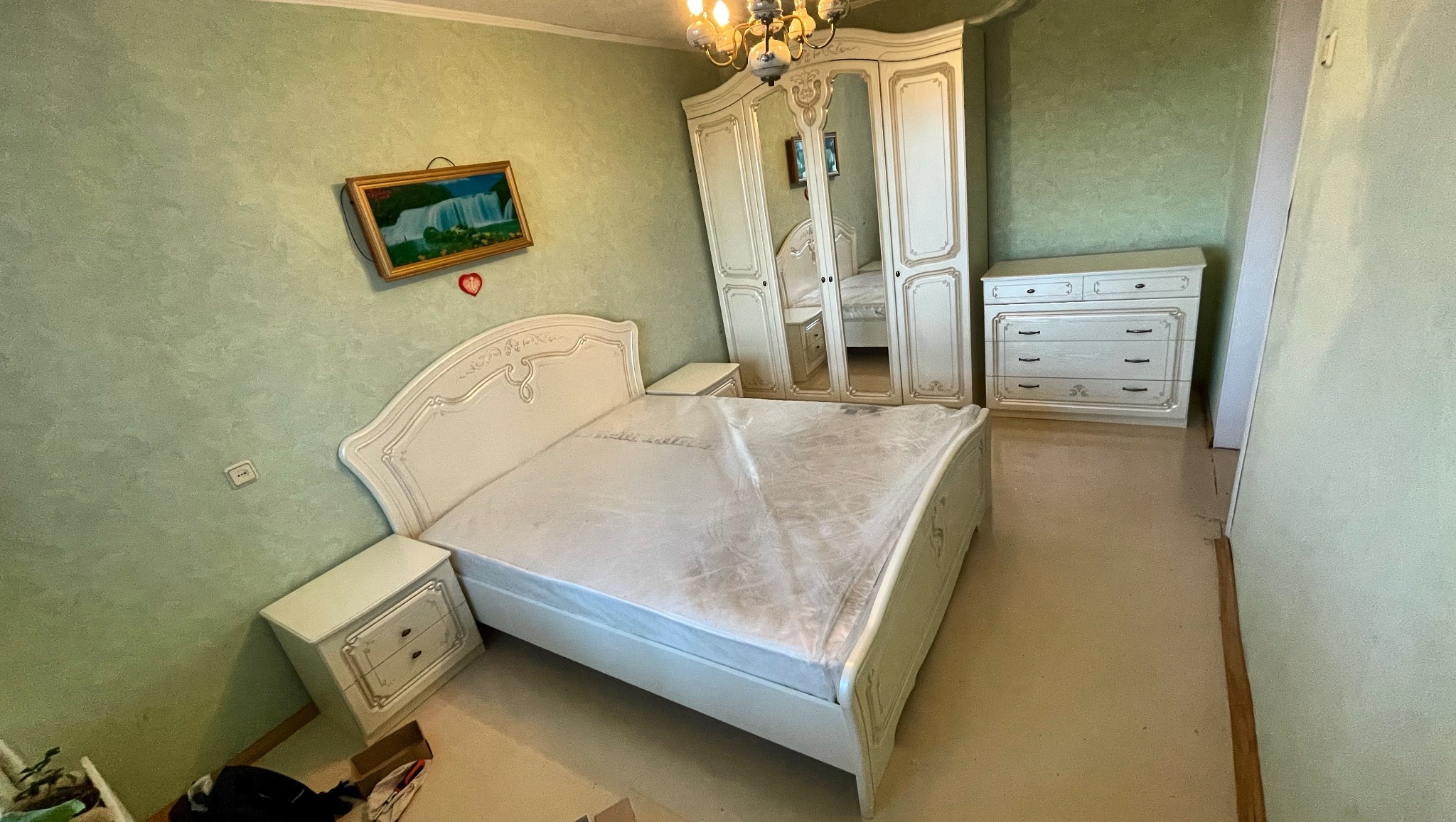 Komplet białych, klasycznych mebli sypialnianych: łóżko, szafka nocna, komoda i szafa z lustrem. Łóżko zabezpieczone folią. Całość na tle zielonej ściany.