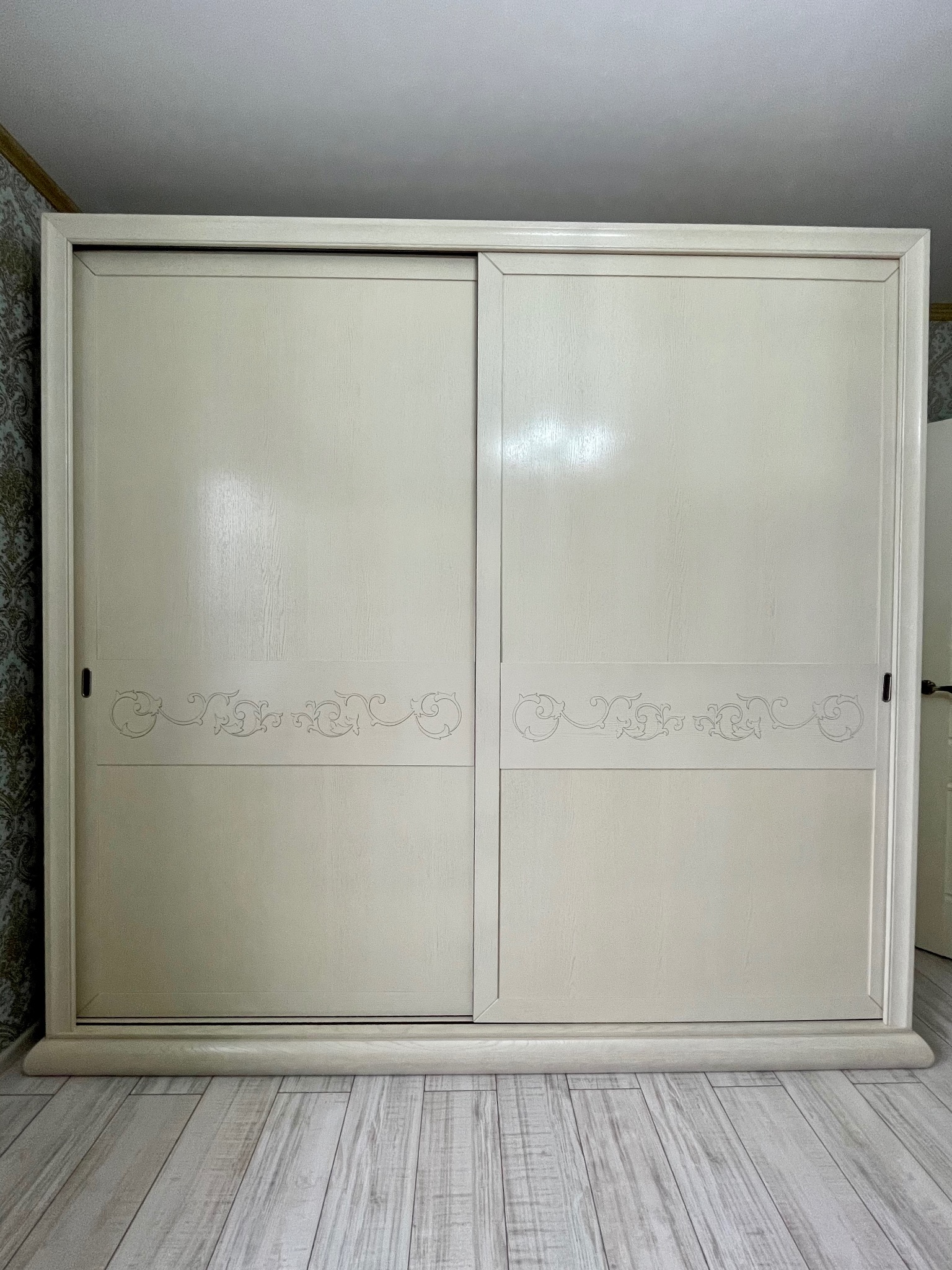 Beżowa szafa przesuwna z subtelnym, dekoracyjnym wzorem na frontach, ustawiona na jasnej podłodze. Widok z przodu, zamknięte drzwi.