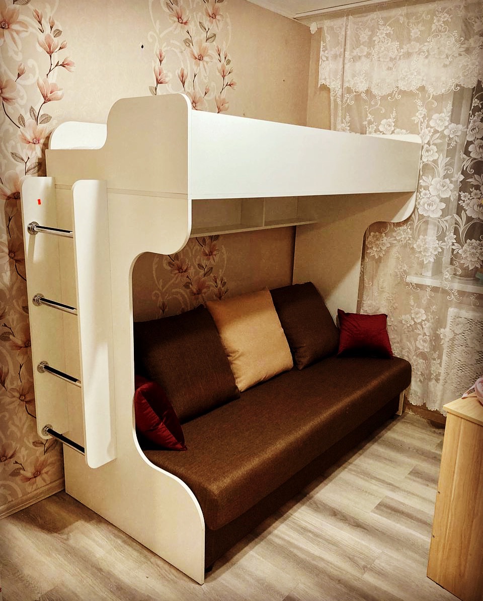 Białe łóżko piętrowe z sofą w pokoju z kwiecistą tapetą. Metalowe drabinki po lewej stronie. Widok na okno z firanką z prawej.