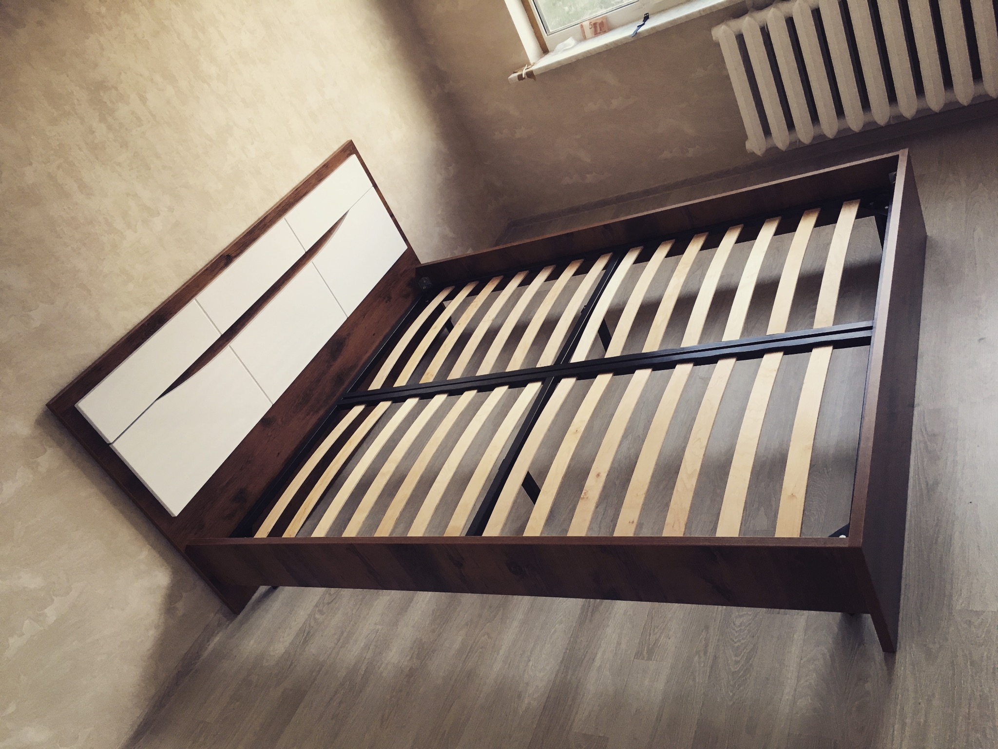 Drewniane łóżko z białym zagłówkiem i elastycznym stelażem listwowym, ustawione w pokoju z grzejnikiem i oknem. Widok z góry, neutralne oświetlenie.