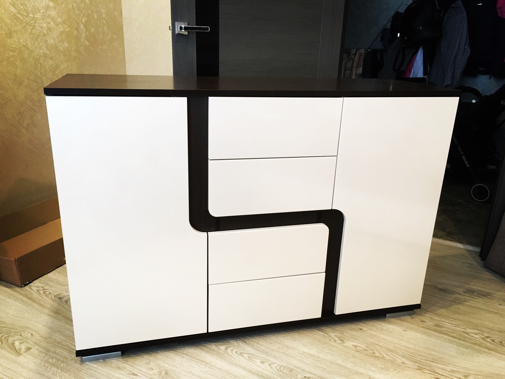 Nowoczesna komoda z białymi frontami i ciemnobrązowymi elementami, stojąca na jasnej podłodze. Widoczne drzwi i szuflady. Charakterystyczny design.