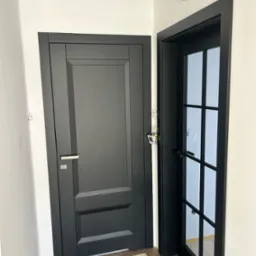 Dwa czarne drzwi: pełne i przeszklone z kratką, zamontowane w białym korytarzu. Widoczne fragmenty podłogi i ścian. Minimalistyczny design.