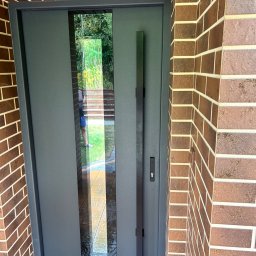 Montaż drzwi i paneli borkowski - Nowoczesne drzwi zewnętrzne w kolorze grafitowym z długim, pionowym uchwytem i szybą, osadzone w murze z cegły klinkierowej. Widok z zewnątrz.