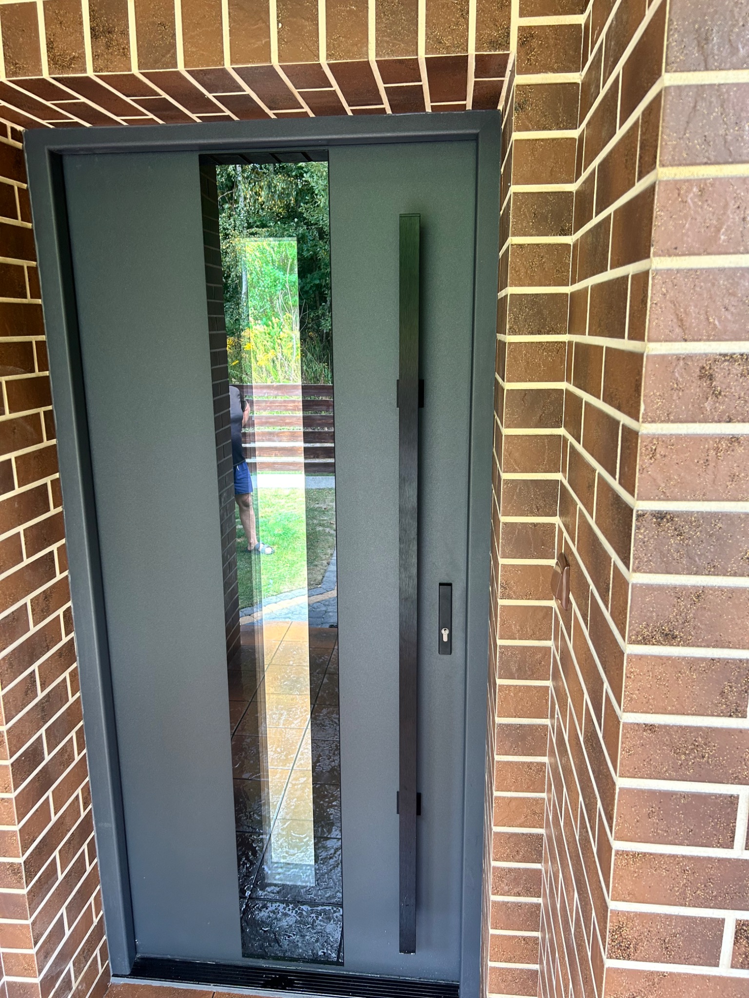 Nowoczesne drzwi zewnętrzne w kolorze grafitowym z długim, pionowym uchwytem i szybą, osadzone w murze z cegły klinkierowej. Widok z zewnątrz.