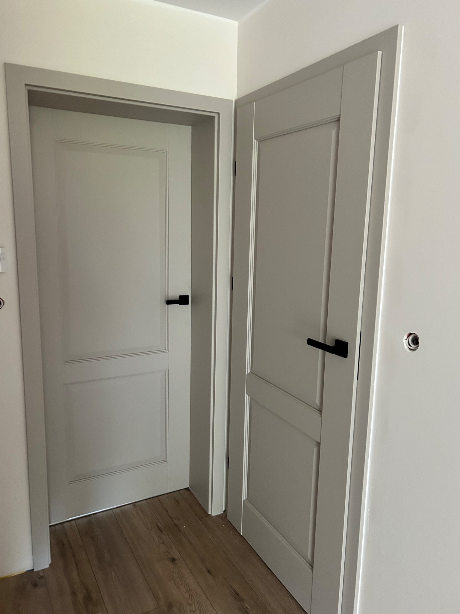 Dwa szare drzwi wewnętrzne z czarnymi klamkami, zamontowane w nowym, minimalistycznym wnętrzu z drewnianą podłogą. Widok z bliska, podkreślający detale wykończenia.