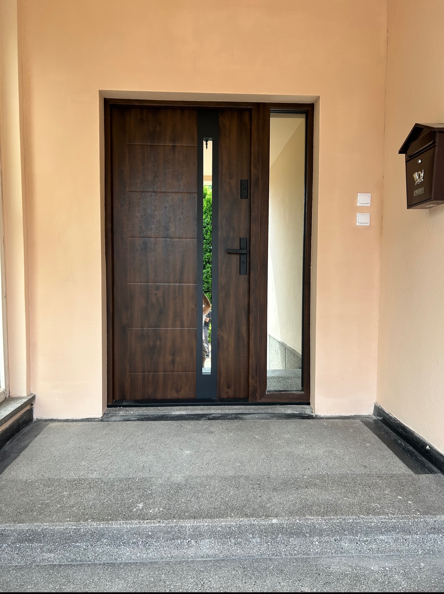 Brązowe drzwi zewnętrzne z szybą, czarny uchwyt i skrzynka pocztowa na beżowej ścianie. Widok z zewnątrz na schody wejściowe.