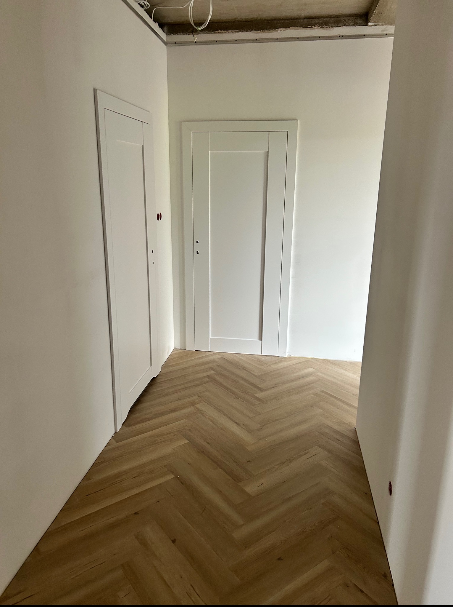 Jasne wnętrze z parkietem w jodełkę i dwoma białymi, prostokątnymi drzwiami wewnętrznymi. Widok na korytarz w trakcie wykańczania.