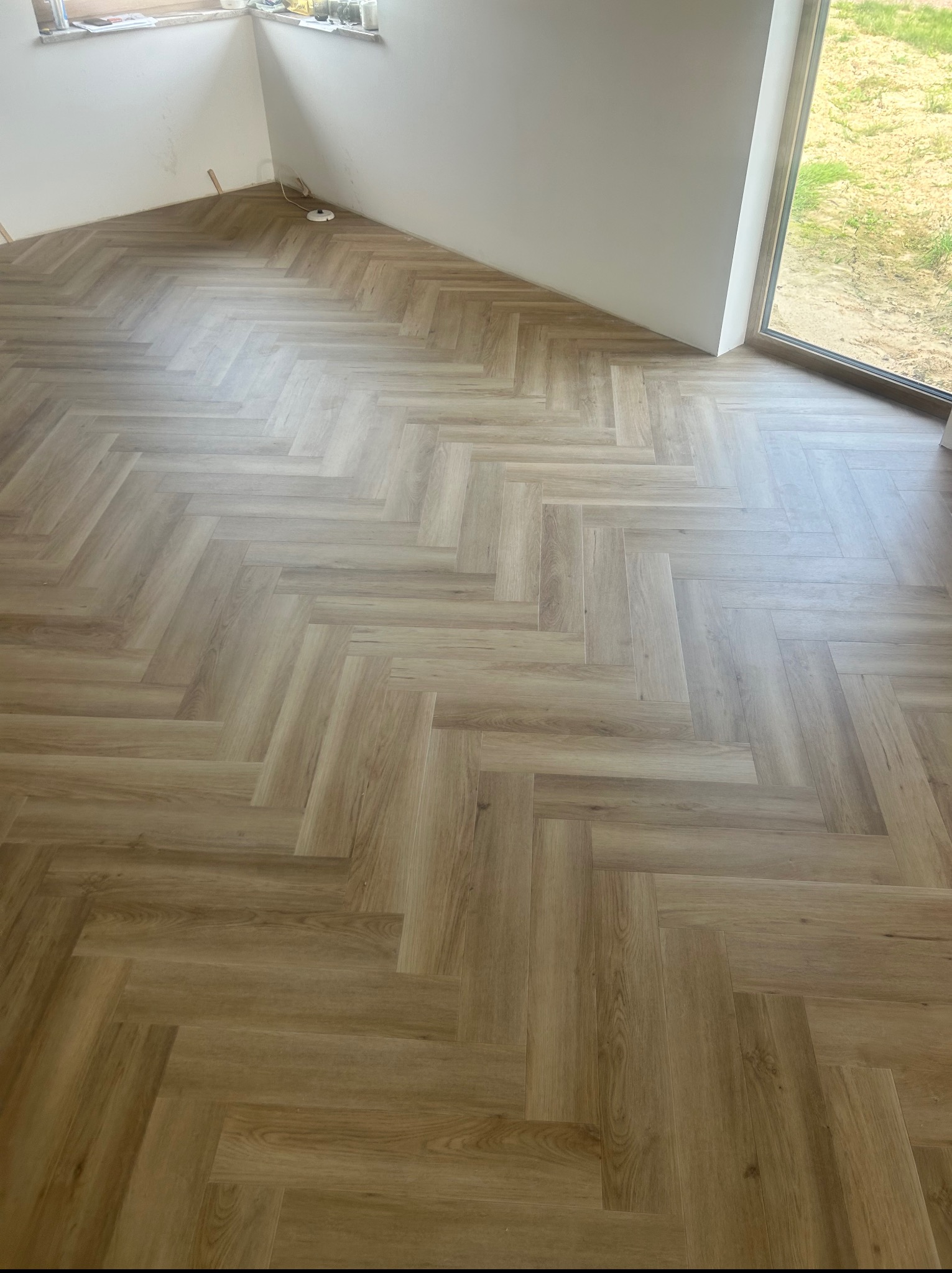 Jodełka z paneli w jasnym odcieniu drewna, ułożona w nowym pomieszczeniu z dużym oknem. Widoczny fragment ściany i listwa przypodłogowa.