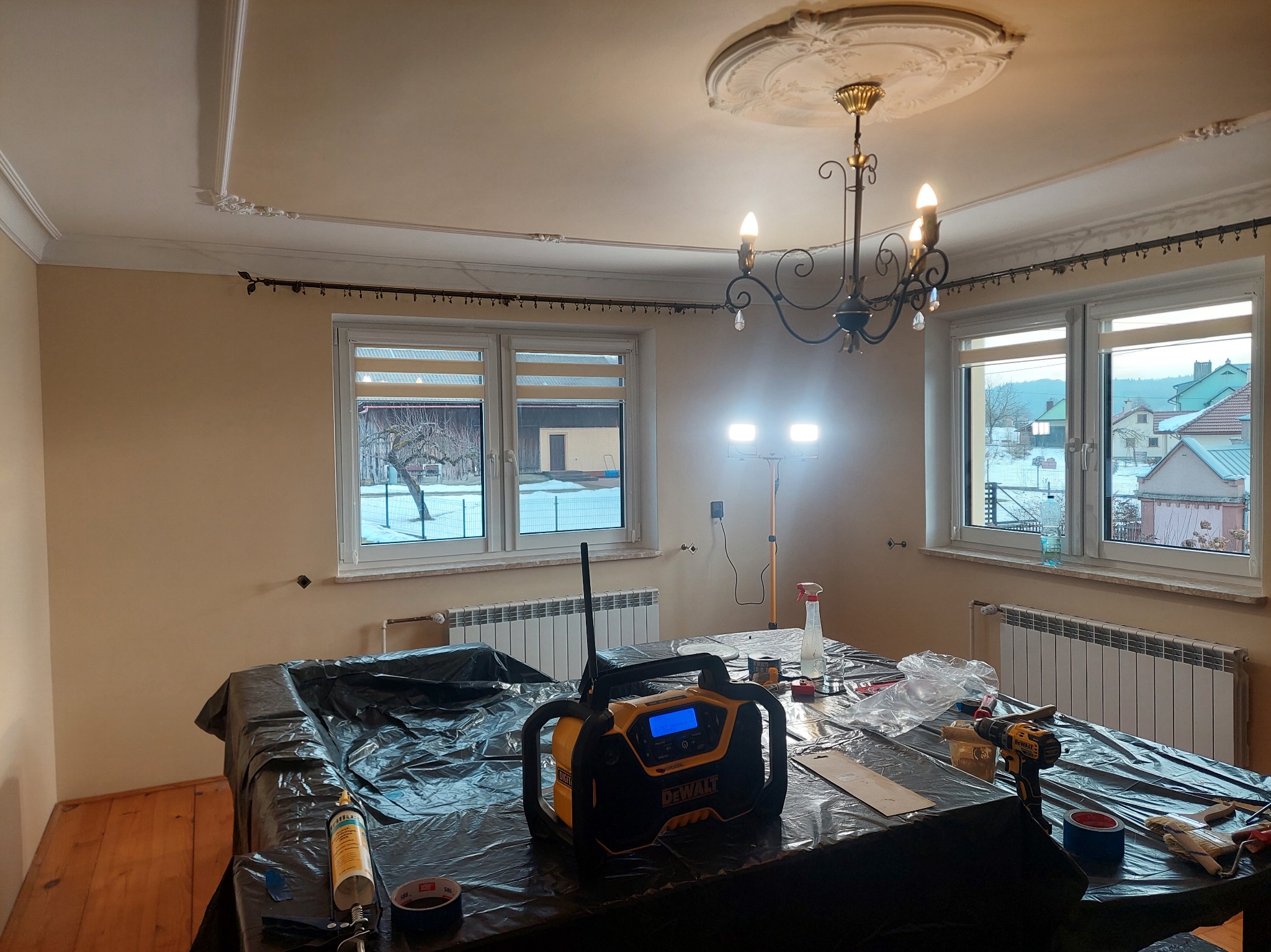 Przygotowania do prac wykończeniowych w jasnym pokoju z oknami. Sprzęt DeWalt na stole przykrytym folią, widoczne narzędzia i akcesoria malarskie. Dekoracyjny żyrandol.
