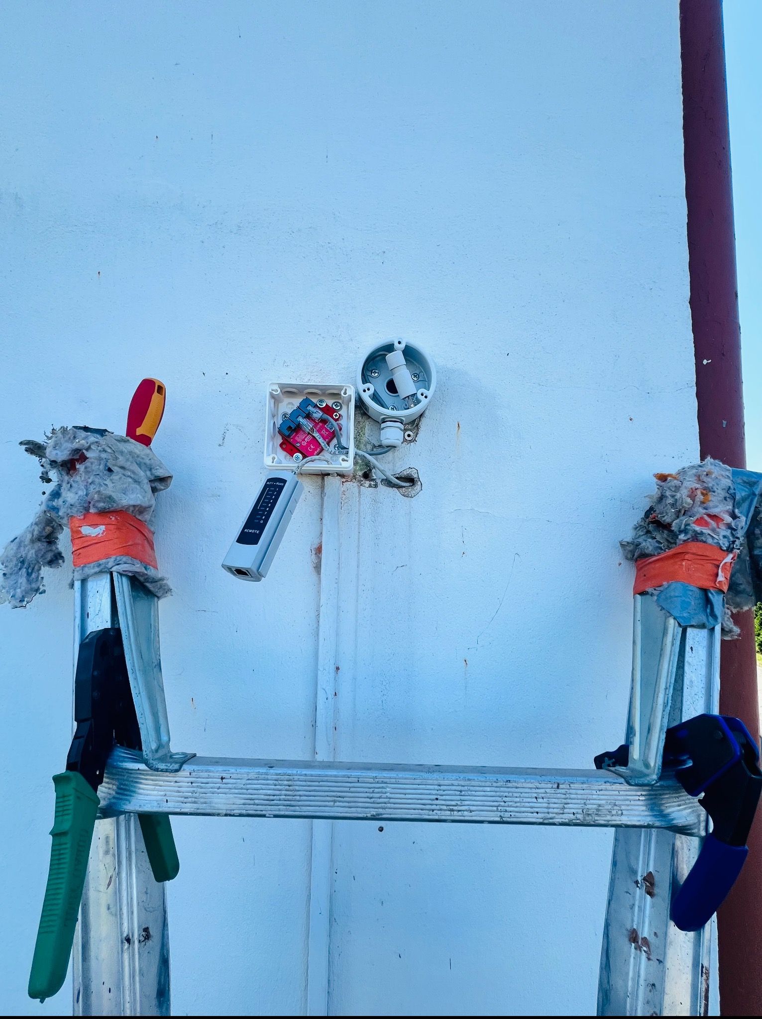 Montaż puszki elektrycznej na białej ścianie z użyciem drabiny i narzędzi, widoczny tester kabli i izolacja. Praca w toku.