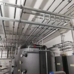 Rozbudowana instalacja grzewcza z kotłem i siecią rur pod sufitem, widoczne elementy mocujące. Profesjonalny montaż instalacji w kotłowni.