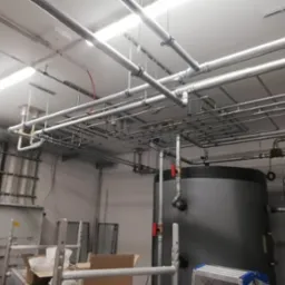 Kompleksowa instalacja rur i przewodów pod sufitem w pomieszczeniu technicznym, widoczny duży zbiornik i skrzynka elektryczna.