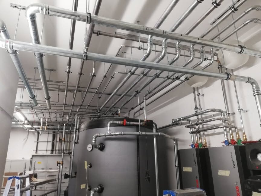 Rozbudowana instalacja grzewcza z kotłem i siecią rur pod sufitem, widoczne elementy mocujące. Profesjonalny montaż instalacji w kotłowni.