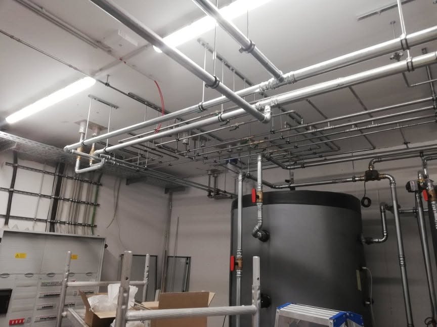 Kompleksowa instalacja rur i przewodów pod sufitem w pomieszczeniu technicznym, widoczny duży zbiornik i skrzynka elektryczna.
