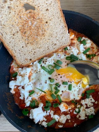 Szakszuka w czarnej patelni z kromką chleba, posypana zielonym szczypiorkiem i serem feta, z widocznym żółtkiem i łyżką. Zdjęcie z bliska.