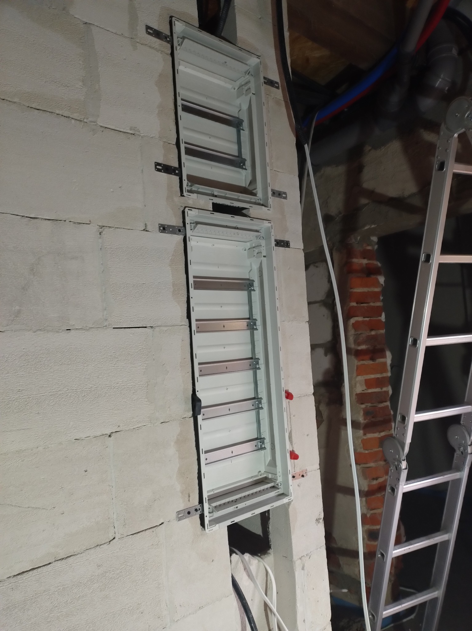 Dwie puste, białe skrzynki elektryczne zamontowane na surowej ścianie z betonu komórkowego. Widoczna drabina i fragment instalacji. Etap montażu rozdzielnicy.