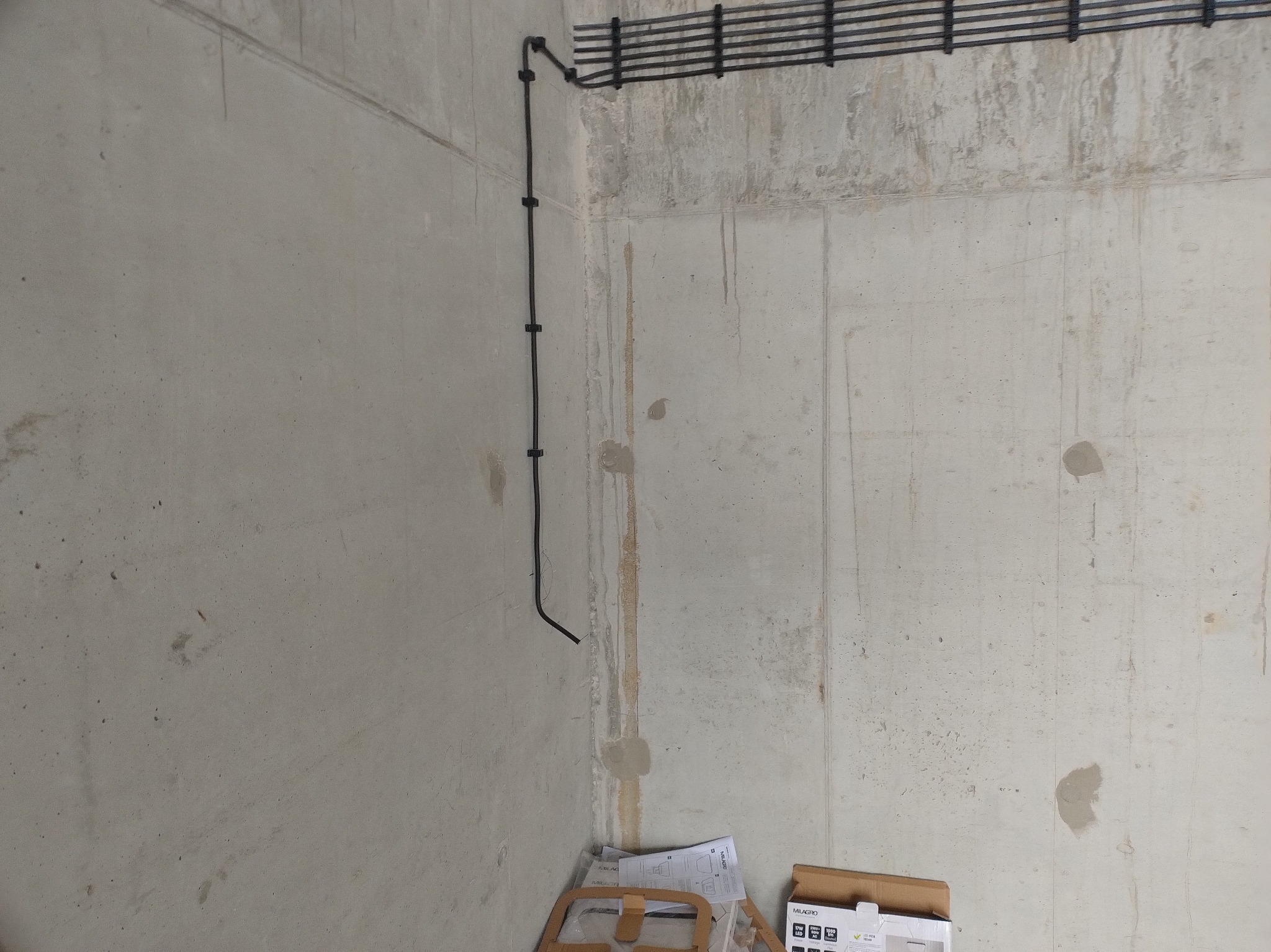 Natynkowa instalacja elektryczna w surowym wnętrzu z betonu. Czarne przewody przymocowane do ściany, widoczna rozdzielnia i osprzęt firmy Simon. Minimalistyczny design.