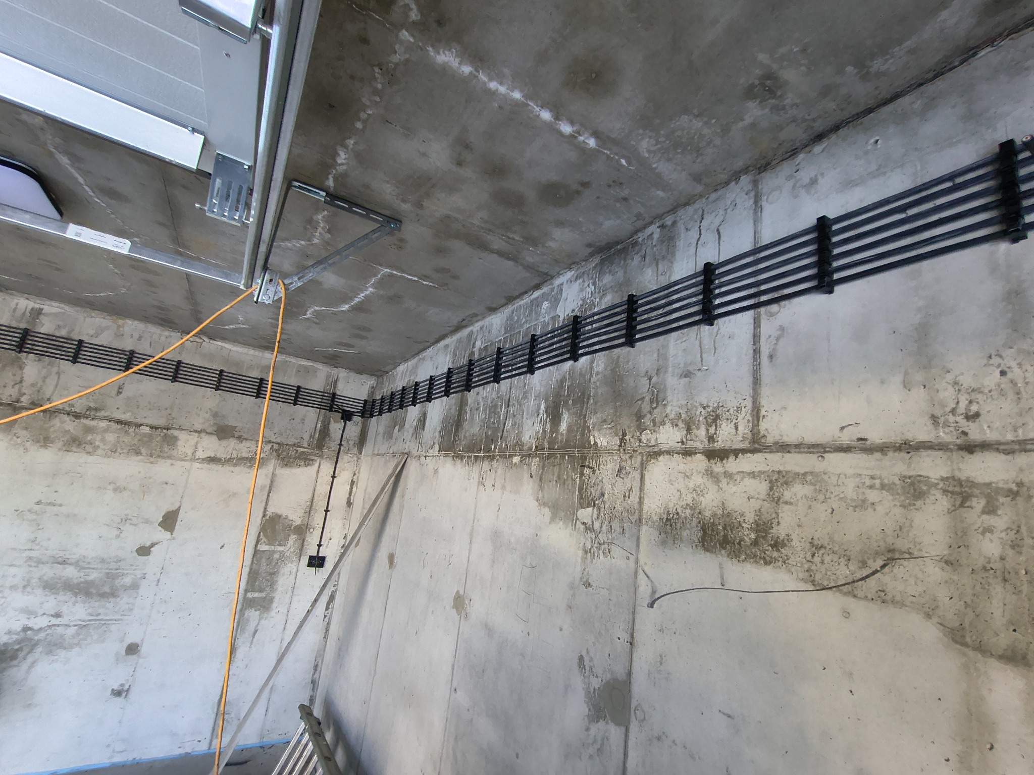 Surowy betonowy sufit z zamocowanymi czarnymi kablami w korytkach i pomarańczowym przewodem, widok z dołu, surowy stan wykończenia.