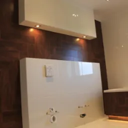 Nowoczesna łazienka z brązową, drewnopodobną okładziną ścienną, białymi płytkami podłogowymi i zabudową geberit z białymi płytkami, widoczne przyłącza wodno-kanalizacyjne oraz górna szafka...