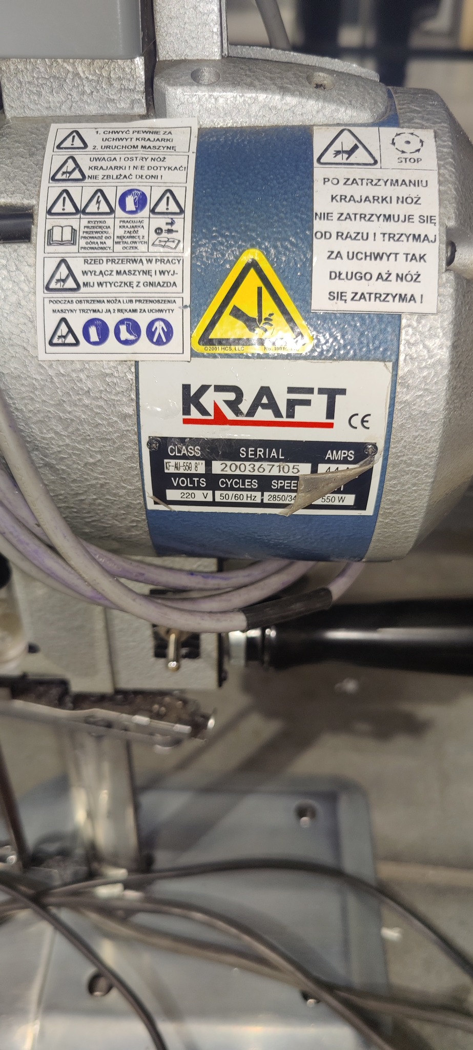 Szczegół maszyny krajarki Kraft z tabliczką znamionową i naklejkami ostrzegawczymi. Widoczne kable i elementy konstrukcyjne maszyny.