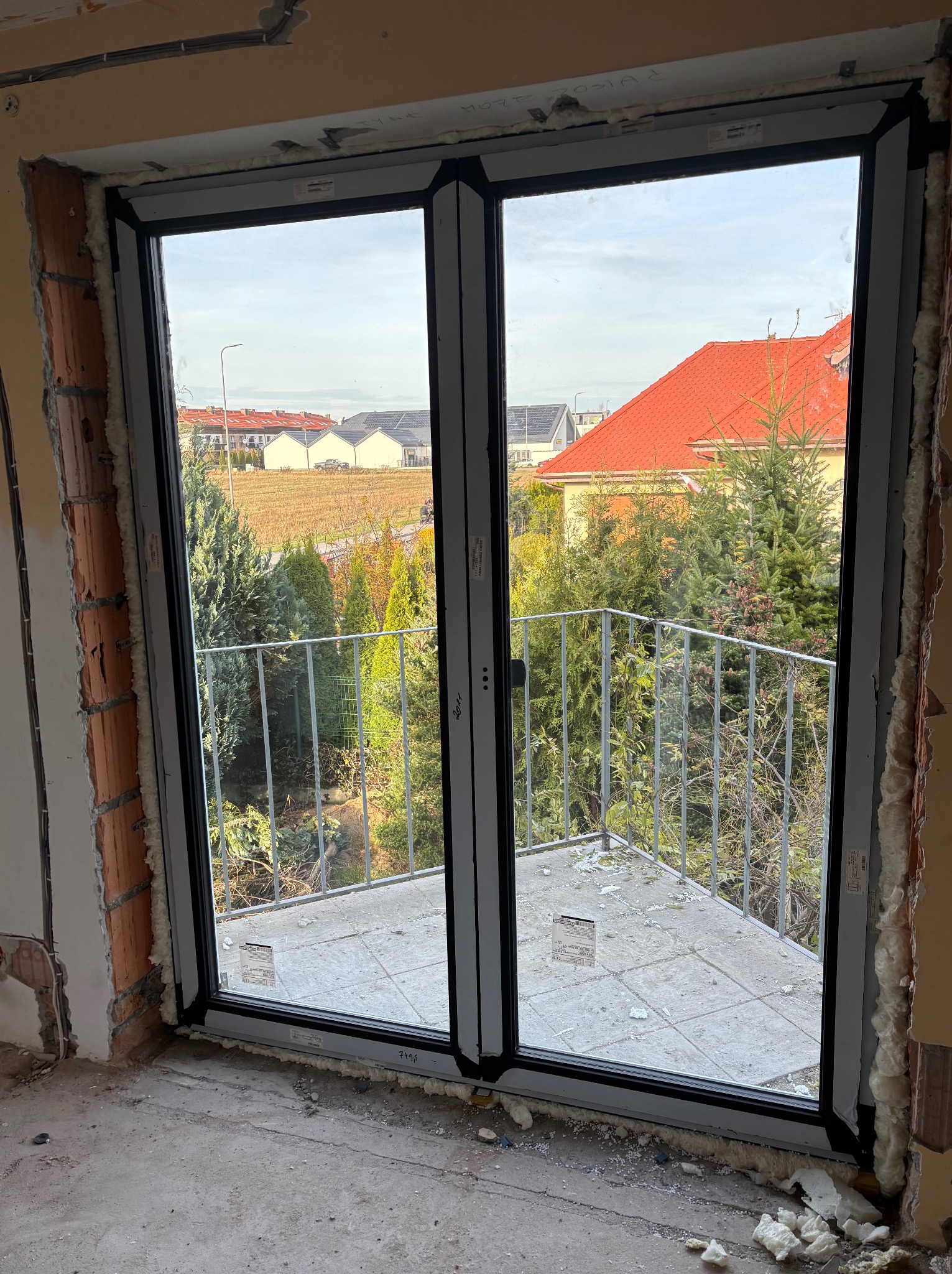 Zamontowane okno balkonowe z widokiem na zewnątrz, pianka montażowa wokół ramy, surowy stan wykończenia wnętrza, widoczny balkon i zabudowa jednorodzinna.