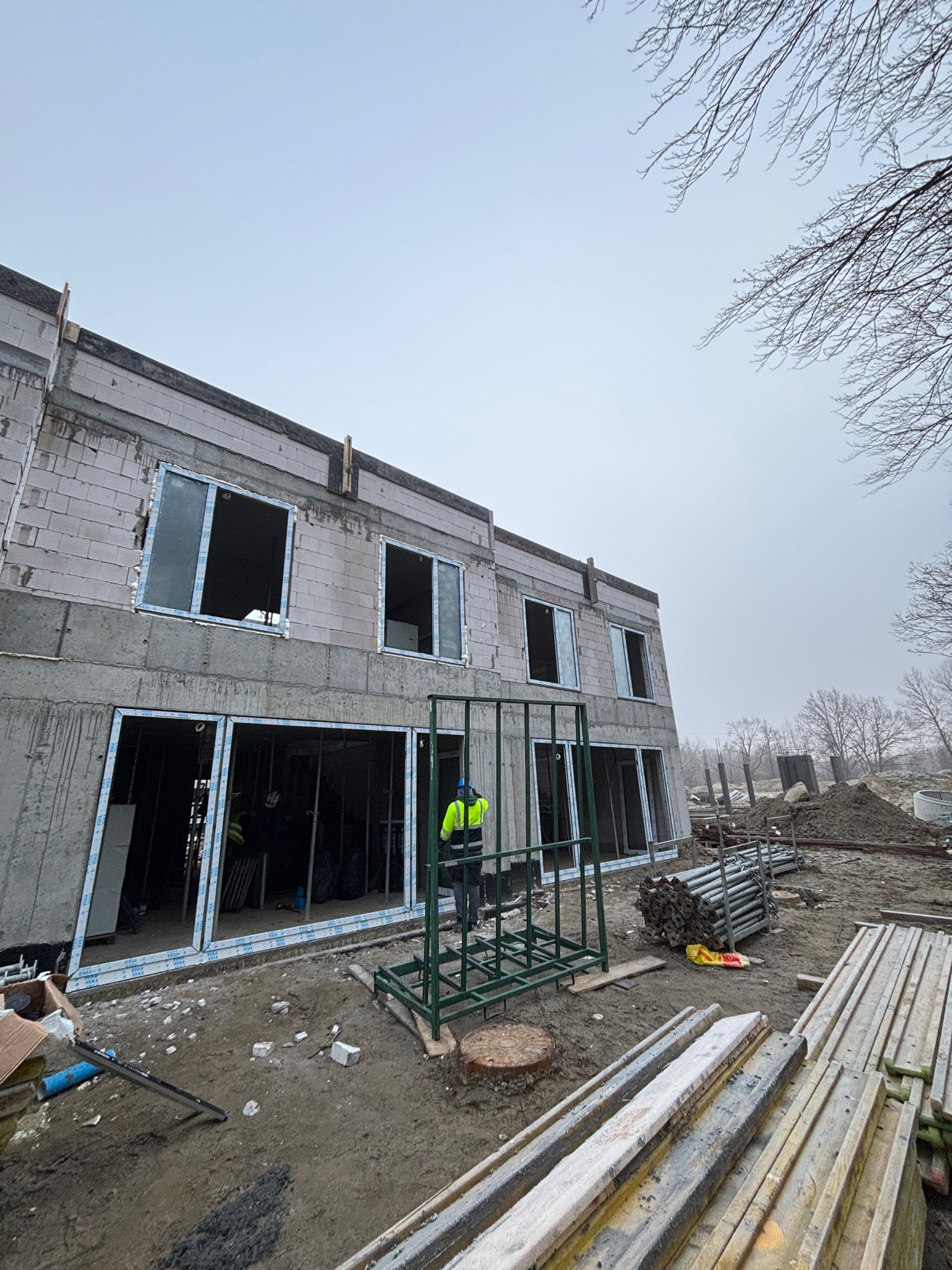 Montaż okien PCV w surowym budynku z betonu komórkowego. Widoczne ramy okienne i pracownik w odblaskowej kamizelce. Surowy stan, plac budowy.