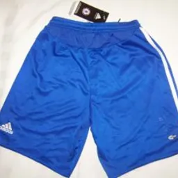 Niebieskie sportowe spodenki Adidas z białymi paskami po bokach, widoczne logo marki w lewym dolnym rogu oraz metka z logo klubu sportowego w górnej części.