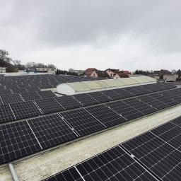 APOLLO SUN SPÓŁKA Z OGRANICZONĄ ODPOWIEDZIALNOŚCIĄ - Rozległa instalacja fotowoltaiczna na dachu budynku, widok z góry, z pracownikami i samochodem dostawczym w tle, pochmurne niebo.