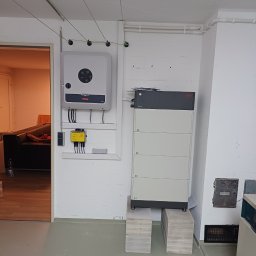 APOLLO SUN SPÓŁKA Z OGRANICZONĄ ODPOWIEDZIALNOŚCIĄ - Instalacja domowa: falownik Fronius i magazyn energii BYD na paletach w piwnicy, widoczne przewody, skrzynka elektryczna i fragment pokoju z kanapą.
