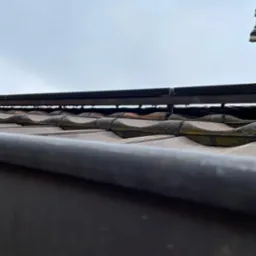 Ujęcie z dołu na ceramiczną dachówkę z metalową konstrukcją montażową i słupem energetycznym w tle, przy pochmurnym niebie.