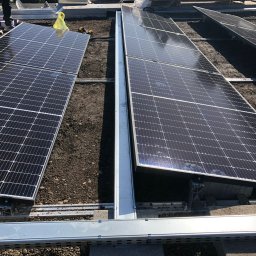 APOLLO SUN SPÓŁKA Z OGRANICZONĄ ODPOWIEDZIALNOŚCIĄ - Montaż paneli fotowoltaicznych na dachu, widoczne aluminiowe konstrukcje wsporcze i częściowo ułożone panele. Prace w toku, widok z góry, oświetlenie dzienne.