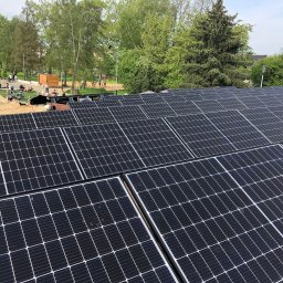 APOLLO SUN SPÓŁKA Z OGRANICZONĄ ODPOWIEDZIALNOŚCIĄ - Montaż paneli fotowoltaicznych na dachu z widokiem na park miejski, gdzie widać osoby odpoczywające na plaży. Instalator w trakcie pracy. Ujęcie z góry.