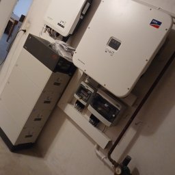 APOLLO SUN SPÓŁKA Z OGRANICZONĄ ODPOWIEDZIALNOŚCIĄ - Instalacja magazynu energii: biała bateria BYD, inwertery SMA, rozdzielnica elektryczna na ścianie w piwnicy. Widoczne rury instalacyjne.