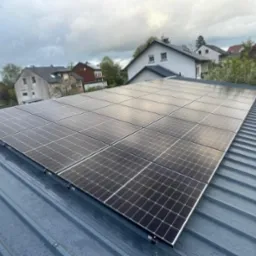 Panel słoneczny na szarym, blaszanym dachu domu jednorodzinnego w pochmurny dzień. Widok z góry, panele ułożone równo, w tle zabudowa mieszkalna.