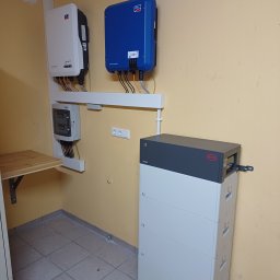 APOLLO SUN SPÓŁKA Z OGRANICZONĄ ODPOWIEDZIALNOŚCIĄ - Kompletny system magazynowania energii: biały akumulator BYD, inwertery SMA (biały i niebieski) i rozdzielnia elektryczna w pomieszczeniu z żółtymi ścianami i płytkami.