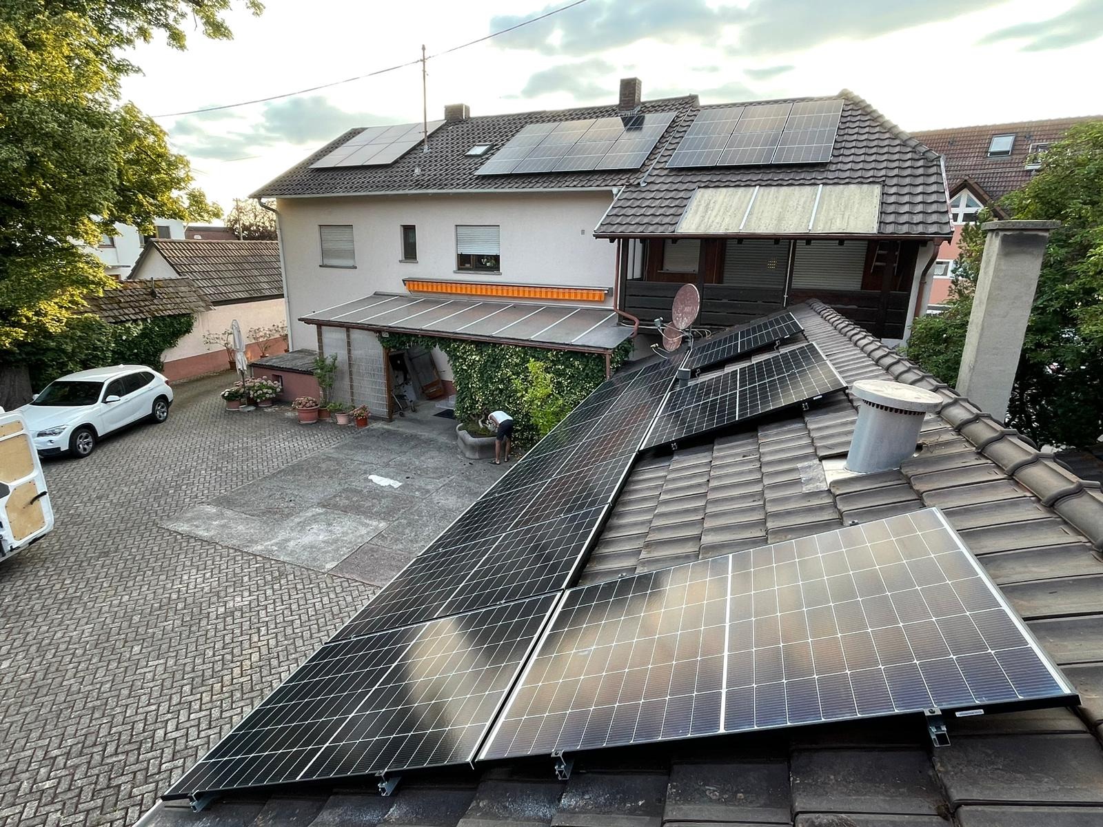 Instalacja fotowoltaiczna na dachu domu jednorodzinnego z brązową dachówką. Widoczny komin, antena satelitarna i biały samochód na podjeździe.