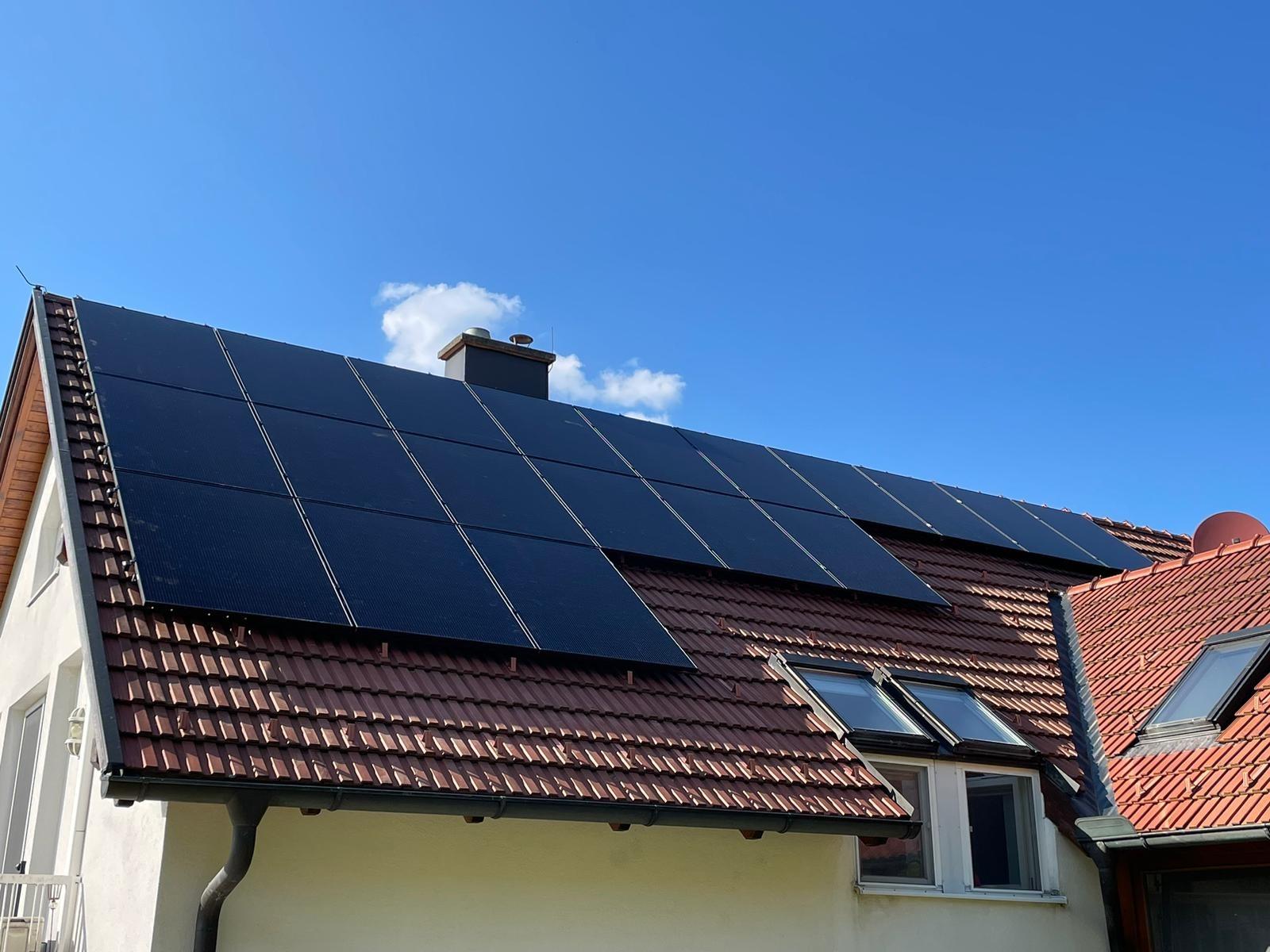 Panele fotowoltaiczne na dachu domu jednorodzinnego z czerwoną dachówką, widoczne okna dachowe i komin. Błękitne niebo w tle.