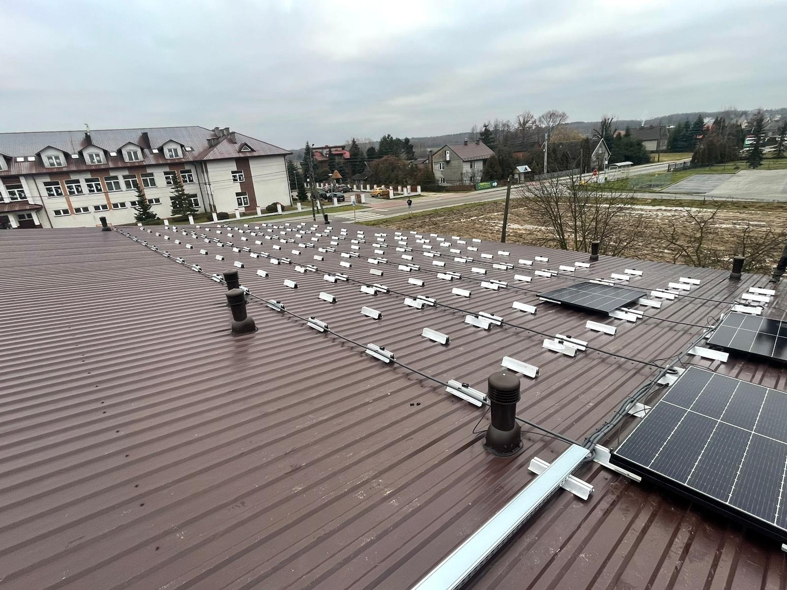 Montaż fotowoltaiki na dachu z blachy trapezowej. Widoczne szyny montażowe i panele w trakcie instalacji. Tło: zabudowa wiejska i pochmurne niebo.