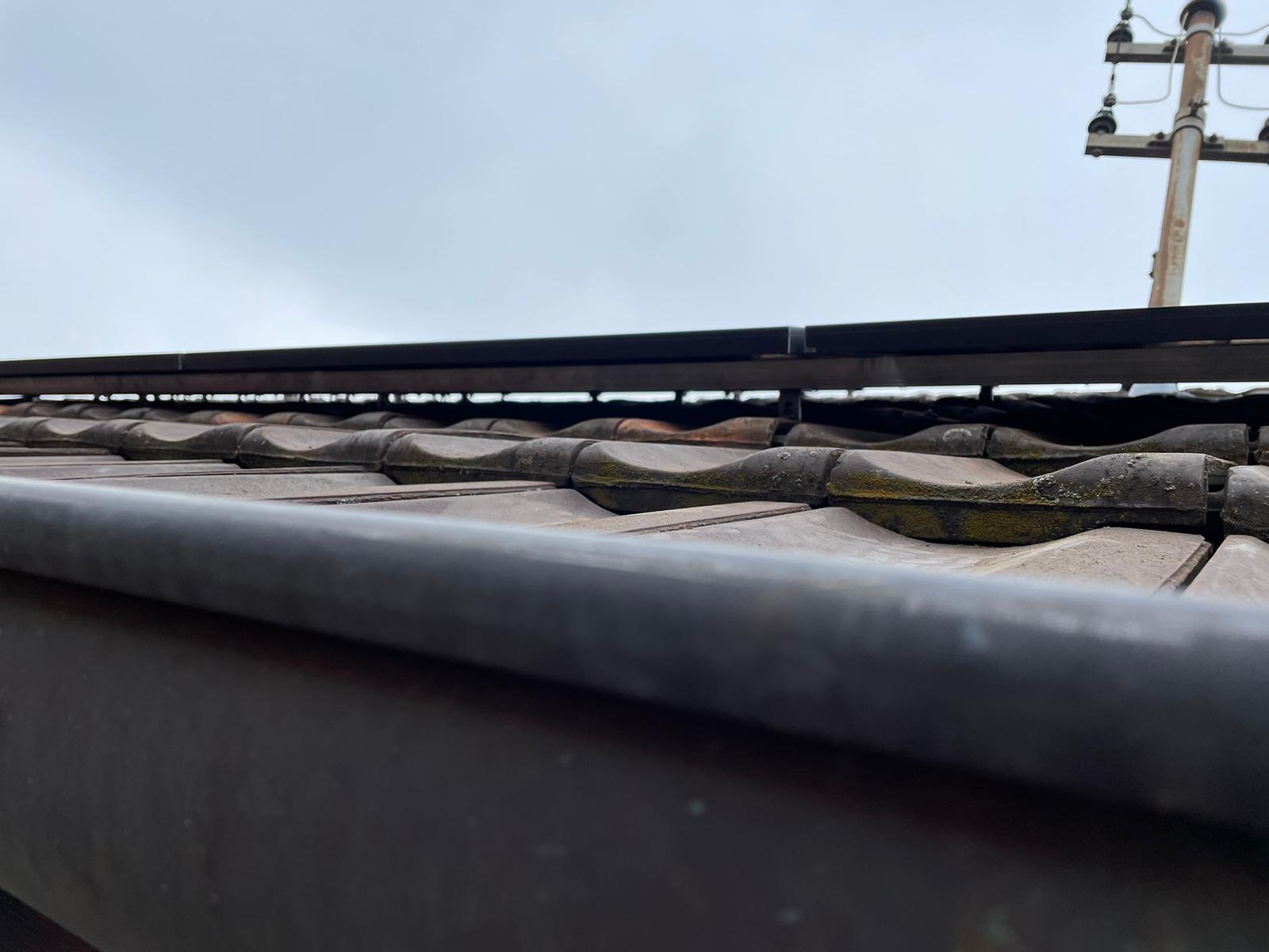 Ujęcie z dołu na ceramiczną dachówkę z metalową konstrukcją montażową i słupem energetycznym w tle, przy pochmurnym niebie.