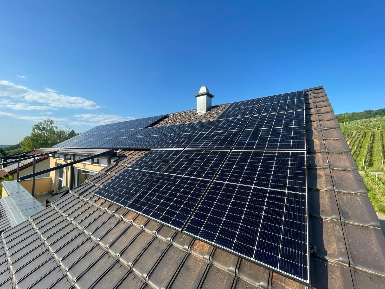 Panele fotowoltaiczne na brązowym dachu domu jednorodzinnego, z kominem i widokiem na winnicę w tle, w słoneczny dzień.