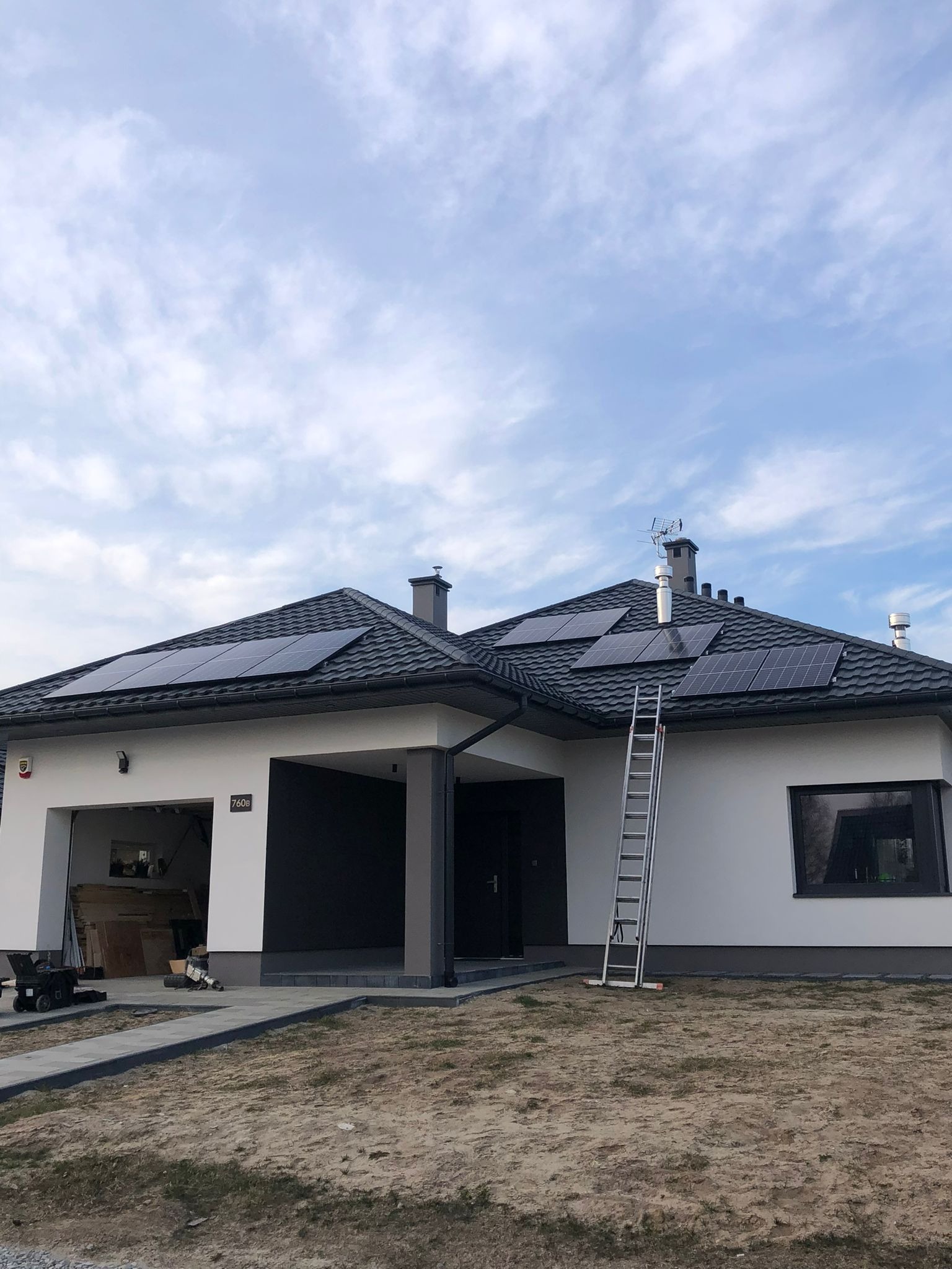 Biały dom z szarą stolarką i panelami słonecznymi na dachu z szarą dachówką. Aluminiowa drabina oparta o ścianę. Częściowo zagospodarowany teren.